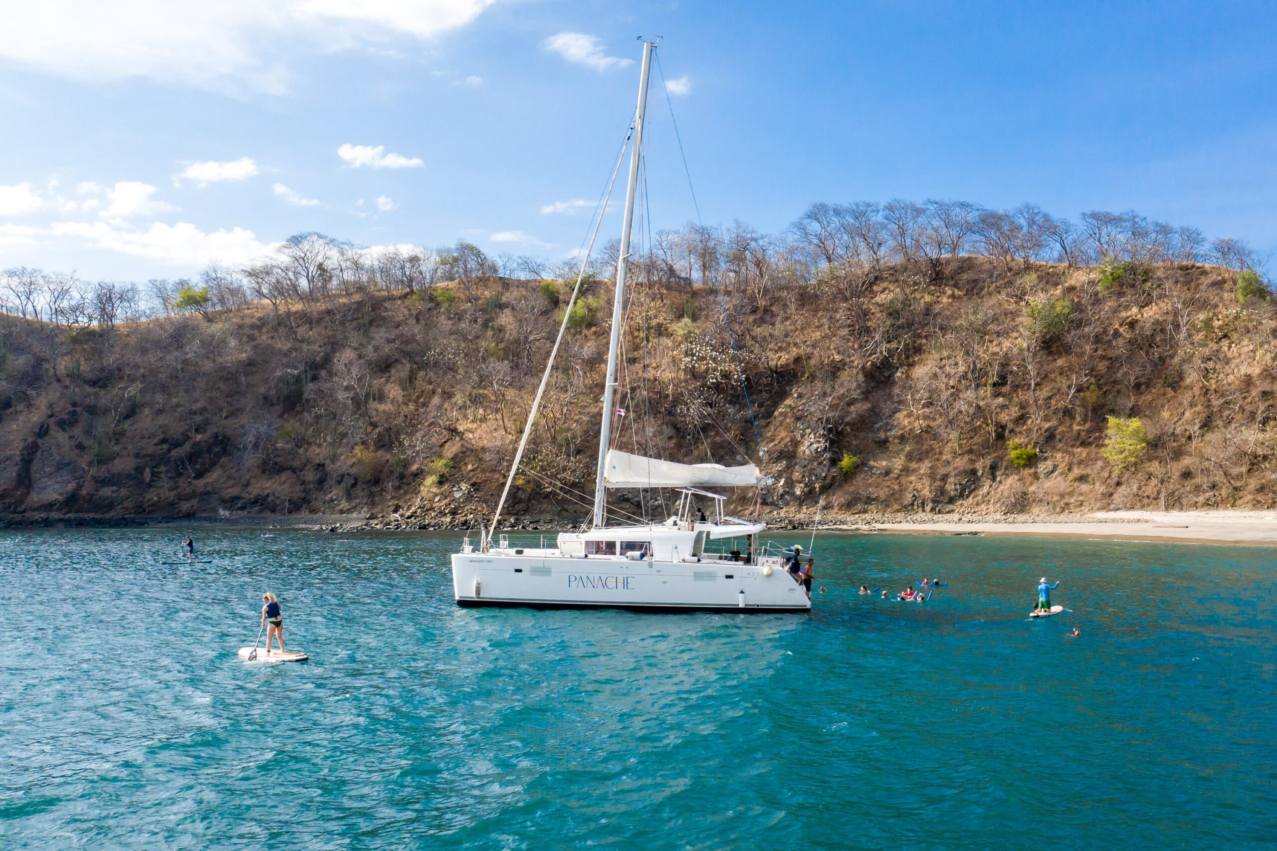 A catamaran anchored near a rocky shoreline, with people swimming and paddleboarding in the turquoise waters under a clear blue sky.
